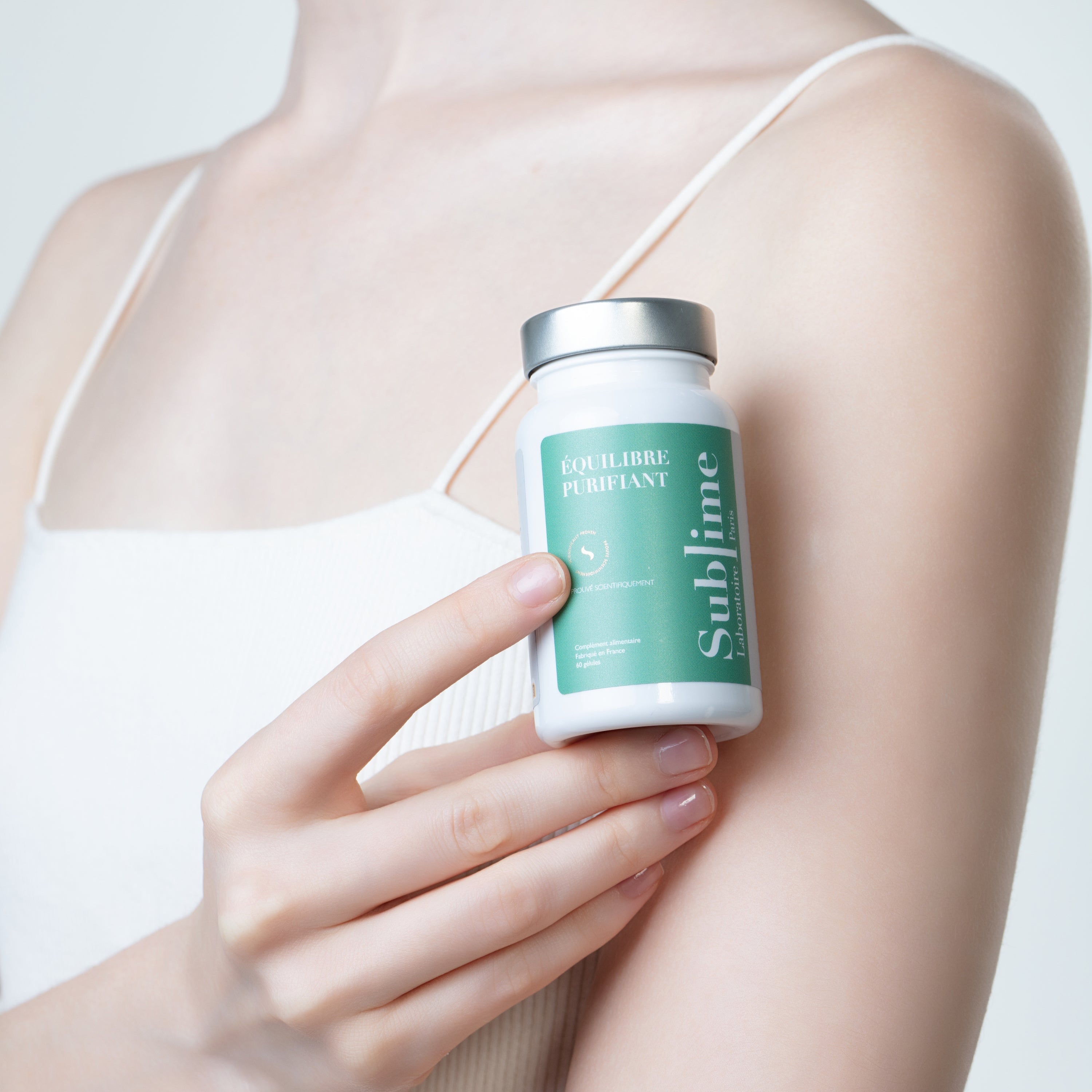 Woman holding Laboratoire Sublime Équilibre Purifiant detox supplement bottle against her arm, French liver and skin detox formula on neutral background