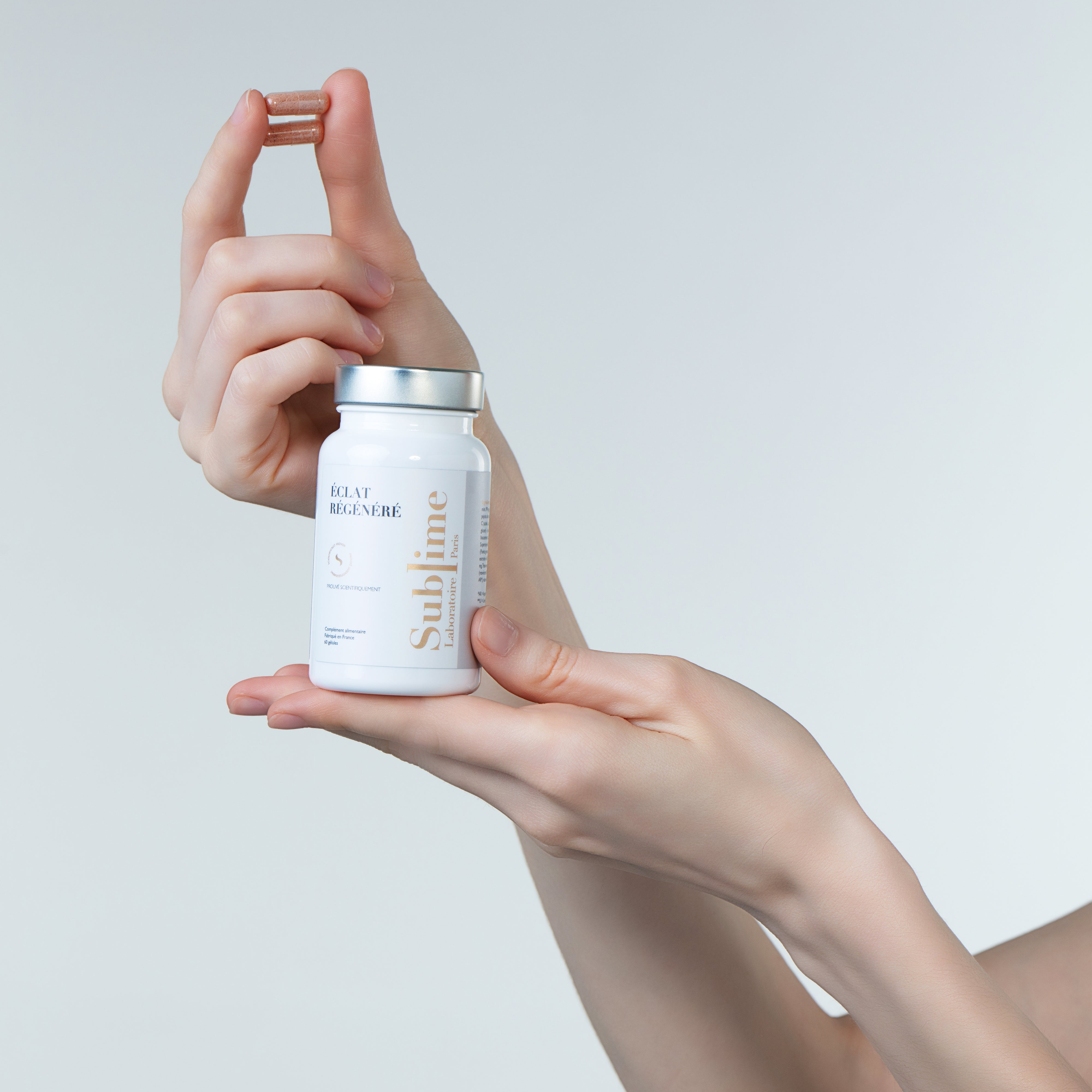 Hand holding Laboratoire Sublime Regenerated Radiance supplement bottle and capsule, French antioxidant and UV-protection supplement, front view on light background