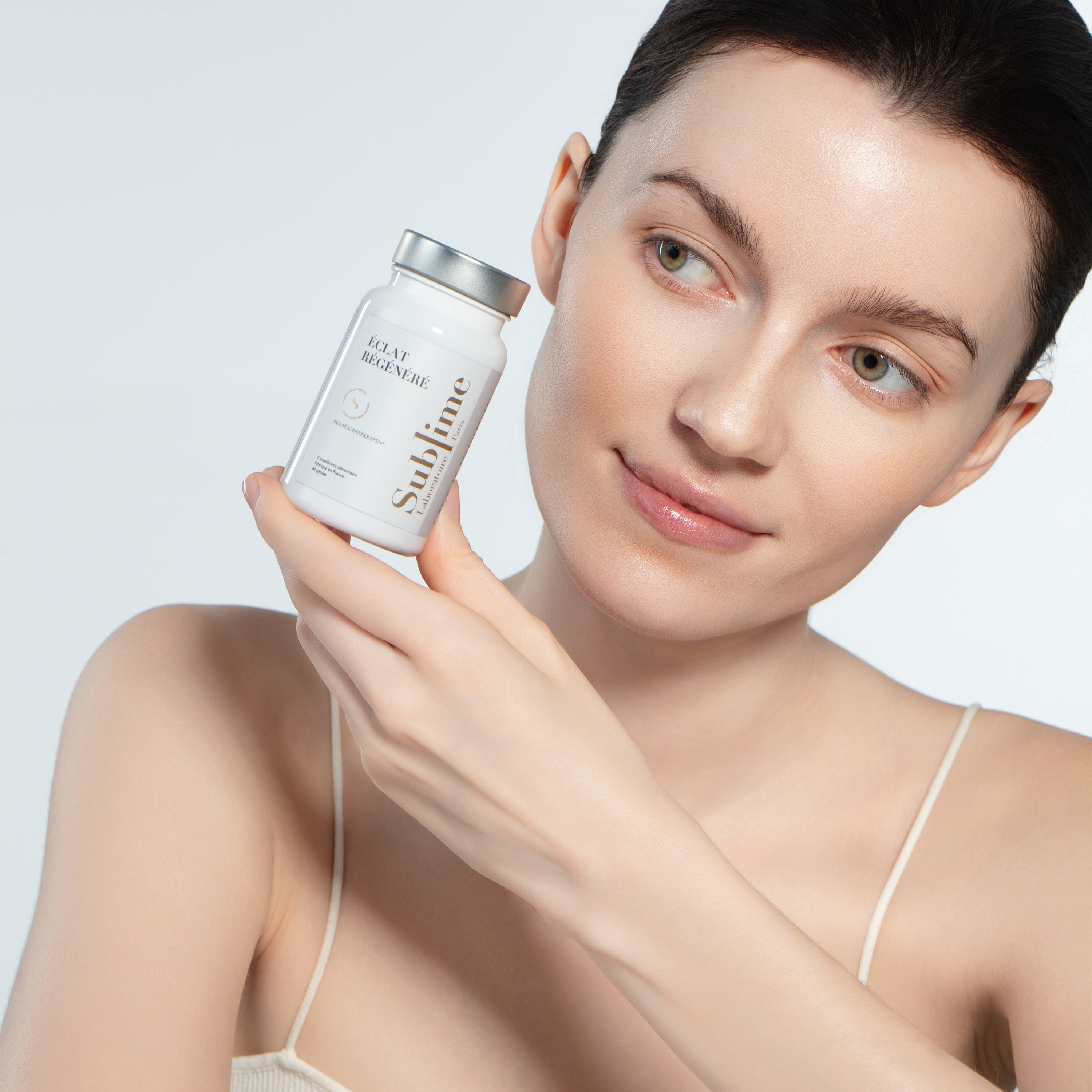 Woman holding Laboratoire Sublime Regenerated Radiance supplement bottle, French antioxidant and UV-protection supplement, front portrait on light background.