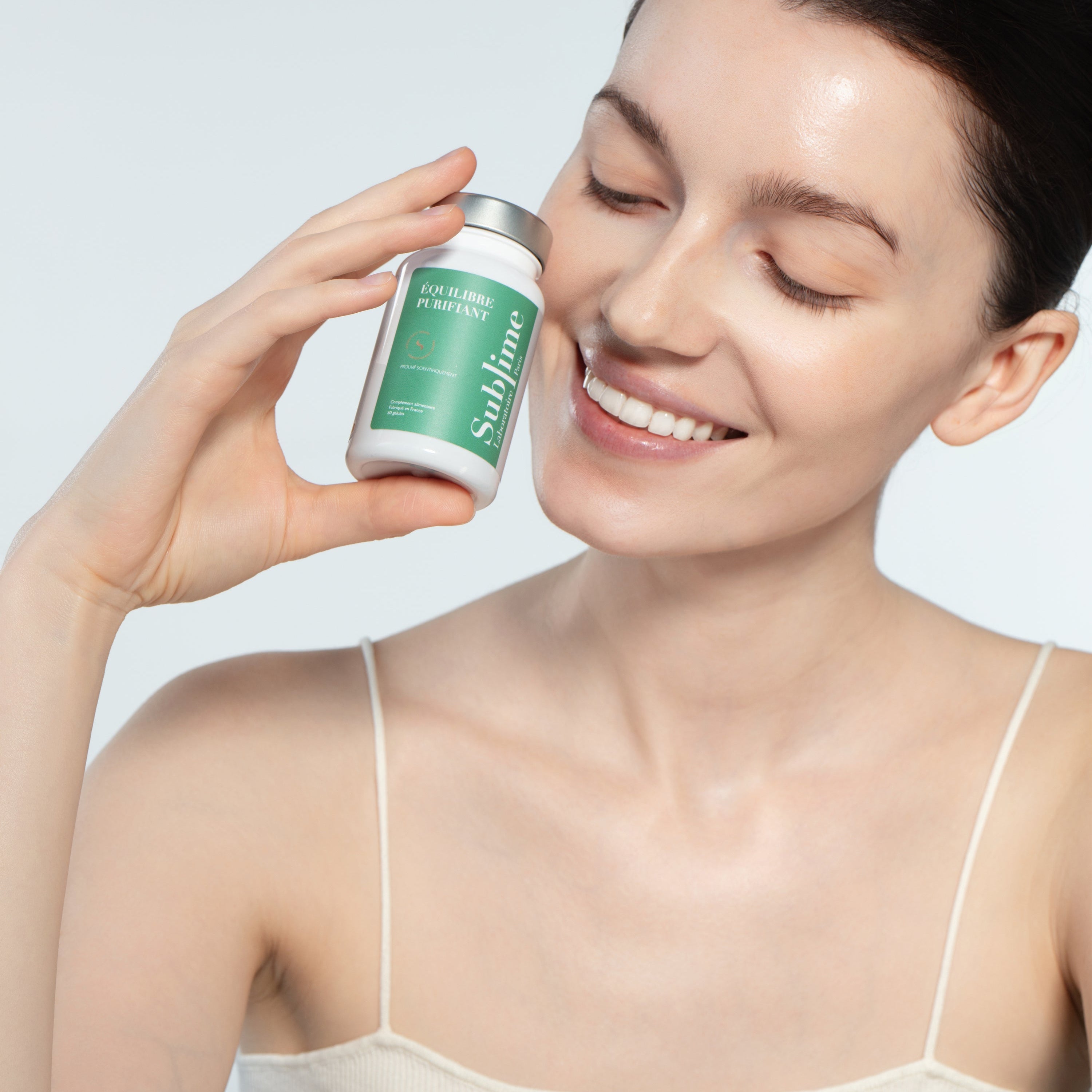 Woman holding Laboratoire Sublime Purifying Balance detox supplement bottle, French liver and skin detox formula, studio portrait on light background